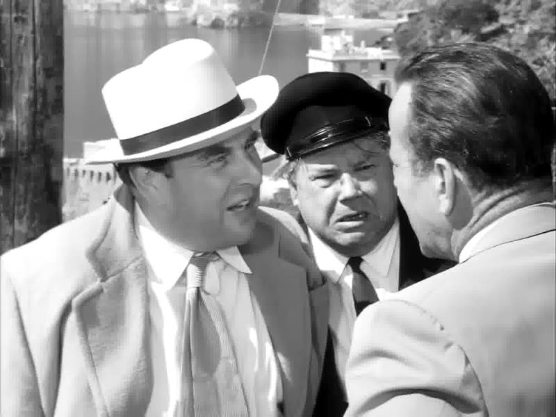 Black-and-white film frame of three men in hats and coats talking closely.